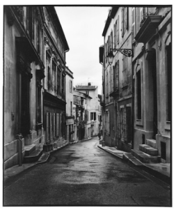 Narrow, empty street lined with old buildings on both sides, photographed in black and white on a cloudy day.