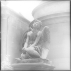 A stone angel statue kneels on a pedestal in front of a blurred, round stone building.
