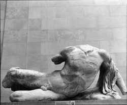 Ancient marble sculpture of a reclining male figure with missing head and limbs, displayed against a plain wall.