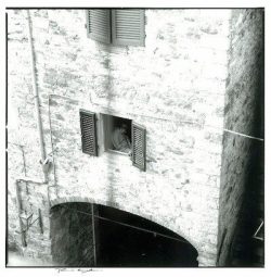 Person looking out from a window with open shutters in a stone building above an archway.