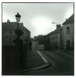 Black and white photo of a quiet street with old buildings, a streetlamp, and a few people walking in the distance.