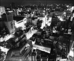 A cityscape at night with densely packed buildings illuminated by artificial lights, viewed from above in black and white.