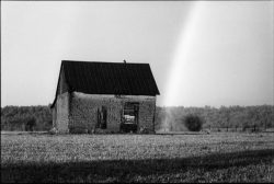 A small, old stone house stands in an open field with a faint rainbow in the sky behind it.