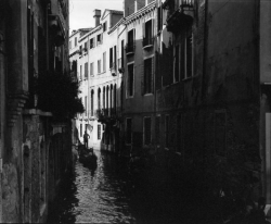 A gondola moves through a narrow canal flanked by old buildings in Venice, Italy, in black and white.