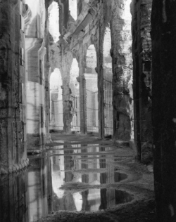 Black and white photo of ancient stone arches reflected in puddles on the floor of a partially ruined building.