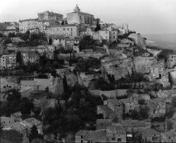 Hillside village with stone buildings and terraced walls, surrounded by trees, under an overcast sky.