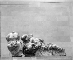 Three headless marble statues from ancient Greece are displayed against a plain stone wall in a museum setting.
