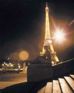 Night view of the illuminated Eiffel Tower with statues and concrete steps visible in the foreground.